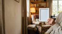 Senior woman with light skin tone holding blank white frame while sitting in comfortable armchair in warm, cozy living room with bookshelf and lamp.