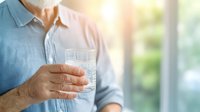 Senior Caucasian man holding glass of pure water in morning sunlight, promoting healthy hydration and wellness lifestyle against blurred window background.