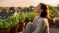 Woman with long dark hair and light skin tone sitting among potted plants on urban rooftop garden, wearing casual beige sweater, looking upward peacefully during golden hour sunset light.