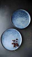 Handcrafted ceramic plates with reactive blue glaze and spotted pattern, one decorated with dried botanical element on dark background. Food photography styling.