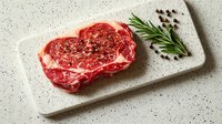 Raw marbled ribeye steak on white stone cutting board with rosemary sprig and peppercorns, ready for cooking on neutral background.