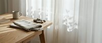 Morning sunlight streams through sheer curtains onto wooden table with newspaper, coffee cup and pastries creating peaceful breakfast scene for lifestyle content.
