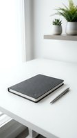 Minimalist workspace with dark gray notebook and pen on white desk, green plants in background creating clean modern office environment for productivity.