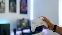 Young adult male with medium brown skin tone holding white gaming controller in modern living room with blurred gaming setup and wall art in background.
