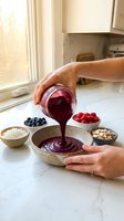 Woman pouring homemade berry smoothie bowl with fresh blueberries and raspberries in bright kitchen for healthy breakfast preparation.