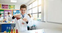 Young Caucasian male student in white lab coat conducting chemistry experiment in bright modern classroom laboratory with colorful educational posters.