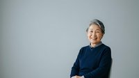 Senior Asian woman with gray hair wearing dark navy sweater, smiling warmly at camera against neutral gray background, suitable for healthcare, wellness, and lifestyle editorial use.