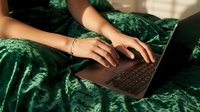 Woman in green dress working on laptop computer from bed in cozy bedroom setting. Remote work lifestyle and digital nomad concept for modern professionals.