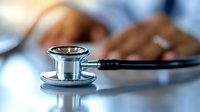 Medical stethoscope on desk with blurred healthcare professional in background working at computer, modern medical consultation concept.