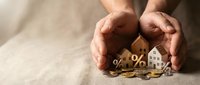 Hands protecting wooden house model with percentage symbols and coins representing home insurance, mortgage rates, and real estate investment protection.