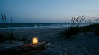 Peaceful beach evening with glowing lantern on sand dunes, sea oats swaying in ocean breeze, twilight sky creating serene coastal atmosphere for relaxation.