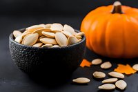Fresh roasted pumpkin seeds in black textured bowl with whole orange pumpkin on dark background, healthy seasonal snack for autumn holidays.