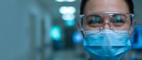 Female healthcare worker wearing protective mask and safety glasses in hospital corridor during medical emergency response and patient care duties.