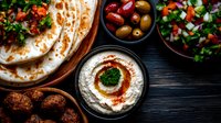 Middle Eastern appetizer spread with hummus, falafel, fresh pita bread, mixed olives and traditional chopped salad on dark wooden table. Mediterranean cuisine.