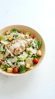 Grilled chicken Caesar salad in wooden bowl with romaine lettuce, cherry tomatoes, croutons, parmesan shavings, and creamy dressing on marble surface.