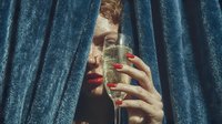Young woman with red manicured nails holding champagne flute behind luxurious blue velvet curtains for celebration and party concepts.