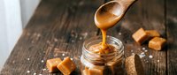 Golden caramel sauce dripping from wooden spoon into glass jar surrounded by caramel candies and sea salt on rustic wooden background for dessert recipes.