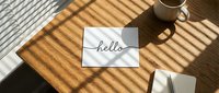 Minimalist workspace flat lay with hello card, coffee mug, and notebook on wooden desk with sunlight shadow pattern.