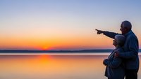 Senior couple enjoying romantic sunset by calm lake water, man pointing at horizon while embracing woman, golden hour reflection creating peaceful moment.