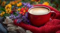 Red coffee cup with creamy foam surrounded by purple wildflowers and orange gerbera daisies on cozy knitted scarf, creating warm autumn mood on rustic wooden surface.