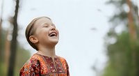 Young girl with light skin tone and blonde hair wearing a red patterned shirt, smiling joyfully outdoors among trees on an overcast day.