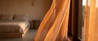 Warm sunlight streams through sheer golden curtains into a minimalist bedroom with wooden furniture and neutral tones, creating a serene morning atmosphere for home decor and lifestyle content.