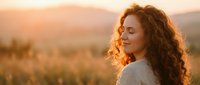 Young woman with curly hair enjoying golden hour sunset in nature, peaceful moment outdoors with warm lighting and mountain landscape background.