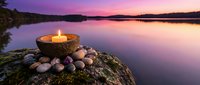 Coconut shell candle with smooth stones on mossy rock by tranquil lake at purple sunset. Meditation wellness spa relaxation mindfulness zen peaceful nature scene.