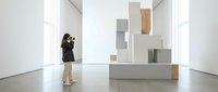 Woman photographer in black jacket and beige pants capturing abstract geometric installation with stacked neutral-toned boxes in minimalist gallery space.