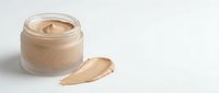 Open glass jar of beige foundation cream with smooth texture and makeup smear sample on white background for cosmetic skincare beauty product advertising.