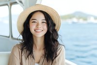 Young Asian woman with long dark wavy hair wearing straw hat smiling broadly while enjoying boat trip on sunny day, ocean and coastline visible in background.