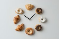 Creative food clock made with assorted pastries and donuts arranged in circle with clock hands on neutral background, representing breakfast time concept.