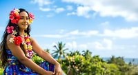 Young woman with long dark hair and medium brown skin tone wearing vibrant floral lei and tropical dress, smiling outdoors with palm trees and blue sky background for travel and cultural themes.