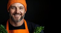 Cheerful Caucasian male chef with beard wearing orange beanie and apron holds fresh herbs against dark background, genuine smile lighting up professional portrait.