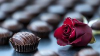 Dark chocolate truffle in brown paper cup with deep red rose on blurred chocolate background, romantic mood for Valentine's Day or special occasion celebration.