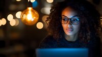 Young African American woman in glasses illuminated by blue laptop light and warm bokeh, working late at night in cozy atmosphere with glowing bulb decoration.