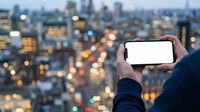 Cropped view of person holding smartphone with blank white screen against illuminated cityscape at dusk, suitable for app mockups and mobile technology marketing.