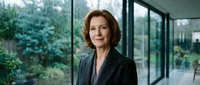 Mature woman with shoulder-length auburn hair and fair complexion wearing dark blazer stands in modern office with floor-to-ceiling windows overlooking green landscape, professional business portrait.