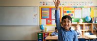 Young Hispanic boy student raising hand enthusiastically in colorful elementary school classroom with educational materials and learning resources.