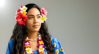 Young woman with long dark hair and South Asian ethnicity wearing vibrant floral lei and blue tropical shirt, adorned with colorful flowers in hair and around neck against neutral background.