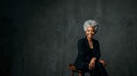 Confident senior woman with gray hair wearing black blazer, seated against dark textured background, professional portrait for business, corporate, and leadership concepts.
