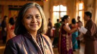 Mature South Asian woman in traditional purple sari smiling warmly at camera during festive celebration with family gathering in background.