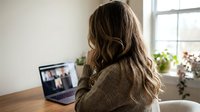 Young woman with long brown hair participating in video conference call from home office workspace with laptop computer and natural window lighting.
