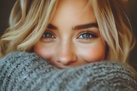 Young blonde woman with blue eyes hiding behind cozy knit sweater, warm intimate portrait with soft lighting for lifestyle and fashion content.
