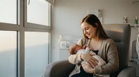 Young Asian mother in cozy sweater breastfeeding newborn baby while sitting in comfortable chair by large window with soft natural light in modern nursery.