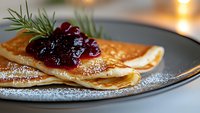 Thin delicate crepes with sweet cranberry sauce and powdered sugar on dark plate, garnished with fresh rosemary sprigs, warm ambient lighting creates cozy mood.
