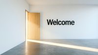 Minimalist white room with wooden door and Welcome sign on wall, sunlight on concrete floor, modern interior for hospitality and business concepts.