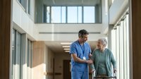 Male nurse in blue scrubs assisting elderly Caucasian woman walking in modern hospital corridor. Healthcare professional providing patient care and support.