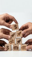Business hands building wooden blocks with startup icons including dollar signs, rockets, gears and team symbols for growth strategy concepts.