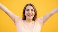 Young woman with shoulder-length brown hair and fair complexion wearing pale pink t-shirt, arms raised in celebration against bright yellow background, expressing joy and success.